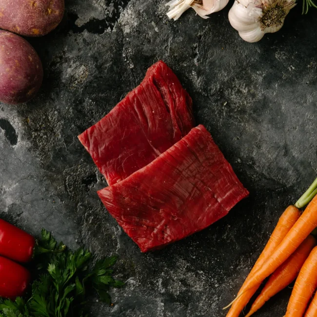 Raw steak, carrots, potatoes, garlic, parsley on dark surface