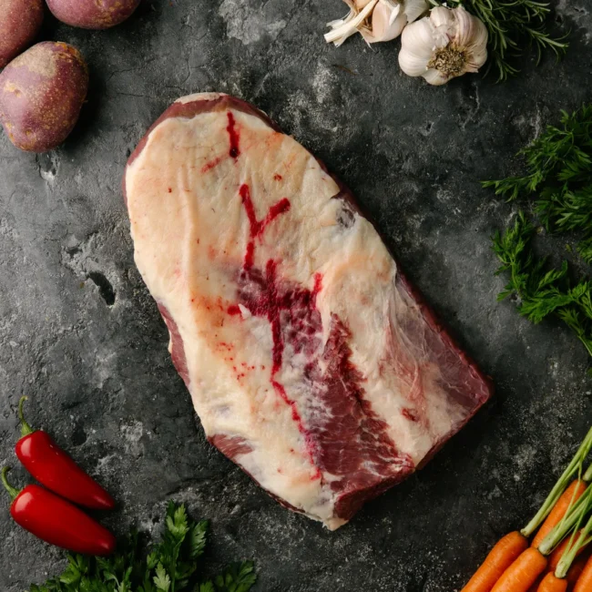 Raw beef brisket with fresh vegetables on slate surface.